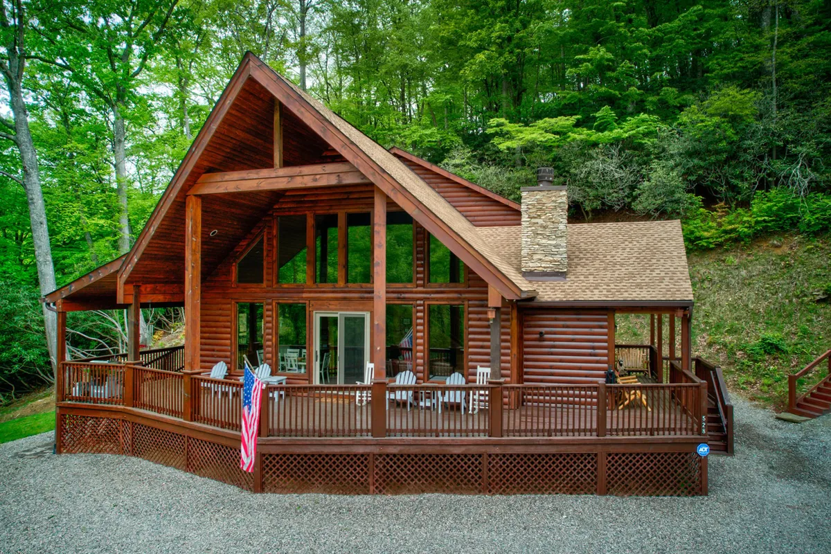 The image features a classic log cabin nestled in lush greenery, with a spacious wooden deck perfect for relaxation. A prominent stone chimney hints at cozy fires inside, while the surrounding trees provide a sense of seclusion and immersion in nature. The welcoming exterior invites guests to imagine sipping coffee at sunrise, hosting gatherings on the deck, or enjoying peaceful evenings under the stars.