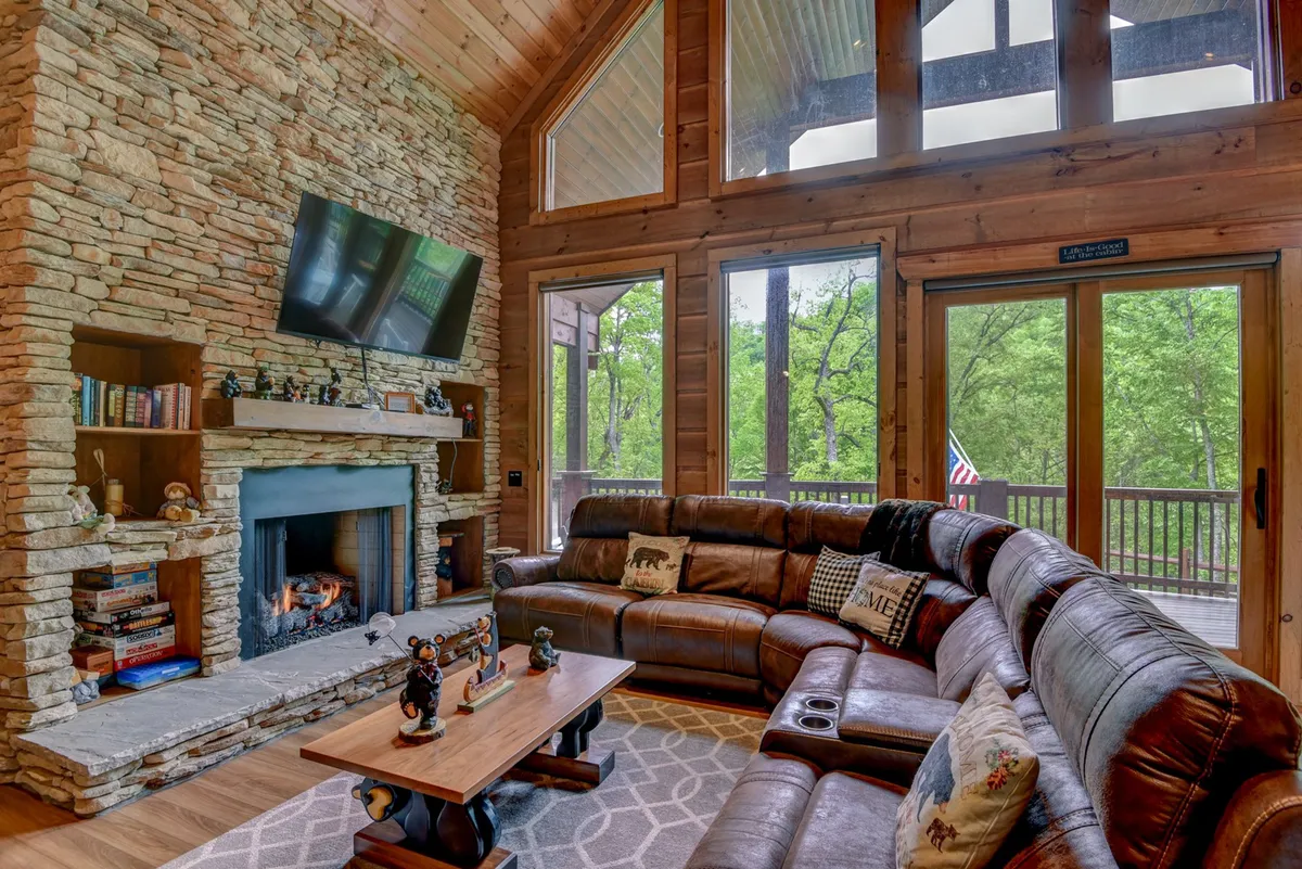 A warm, welcoming living room with a stone fireplace, large windows, a leather sectional sofa, and a wooden coffee table. The space feels inviting and is ideal for cozy winter nights or relaxing after exploring outdoors.