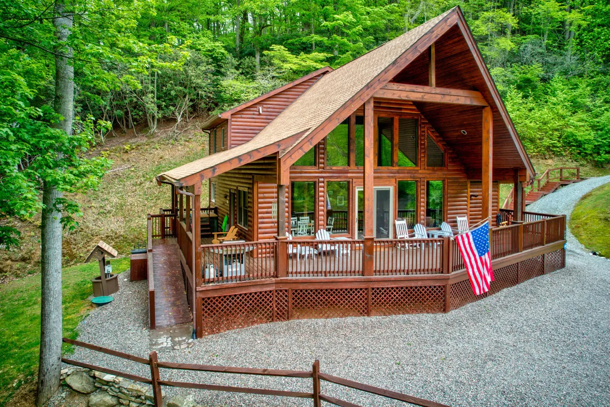 A large wooden deck with expansive glass windows revealing glimpses of the home's inviting interior. The American flag flutters in the breeze, and the deck is clearly suited for gatherings or relaxing while watching sunsets over the mountains.