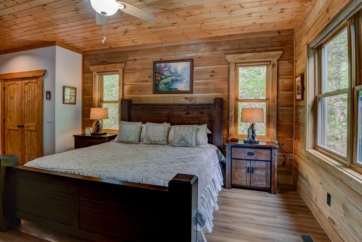A spacious bedroom with a king-size bed dressed in decorative pillows, wooden nightstands with lamps on either side, and tasteful artwork mounted on the wall. Sunlight streams through the windows, creating a serene and restful atmosphere perfect for relaxation.