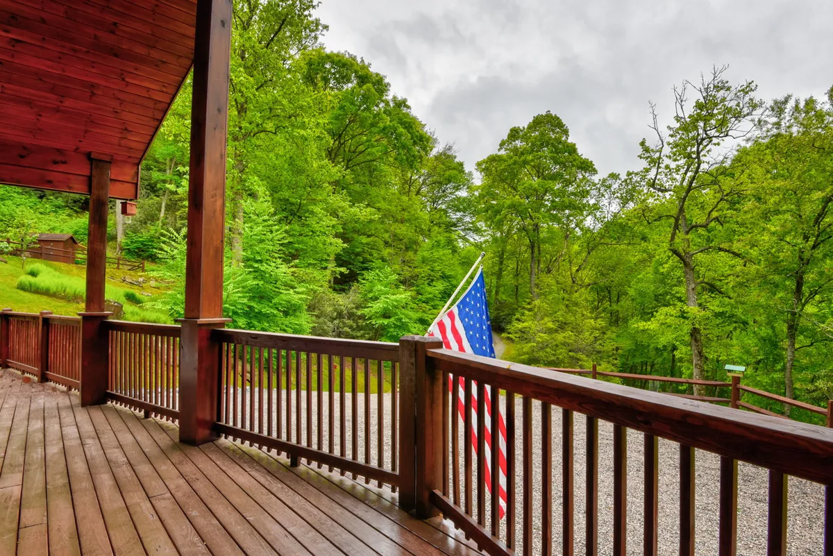 A wooden deck framed by lush greenery and adorned with an American flag, offering tranquil views and an inviting space for outdoor activities and quiet reflection. The natural setting feels both private and expansive, perfect for strolls and moments with pets.