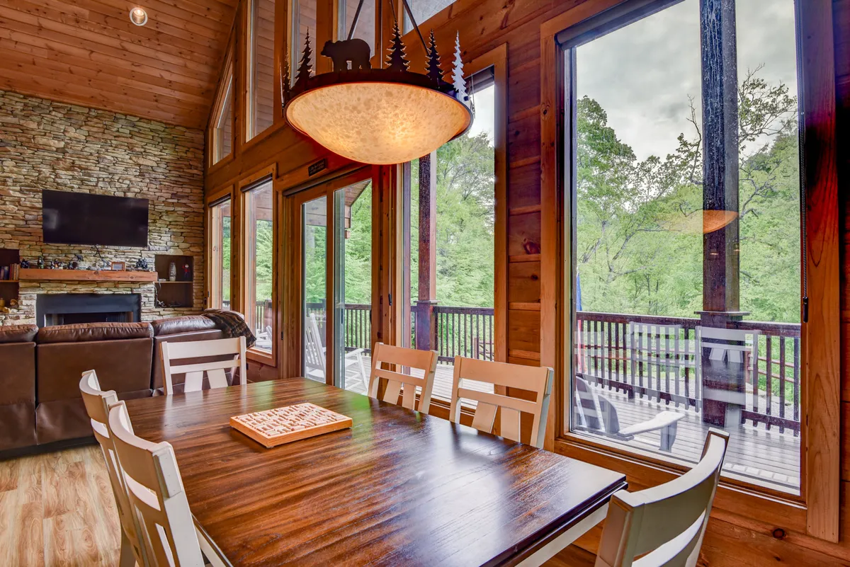 An expansive dining area with a large wooden dining table, a stone fireplace, panoramic windows letting in natural light, and a decorative ceiling light. The space is warm and ideal for both intimate dinners and large family gatherings.