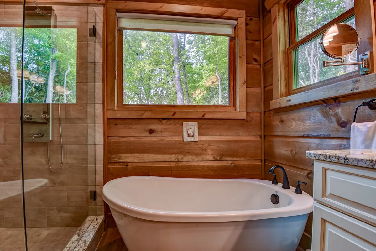 A luxurious bathroom with a freestanding soaking tub positioned by large windows, providing a peaceful spot to relax while gazing out at surrounding trees. The space is elegant and designed for indulgence, reflecting upscale amenities.