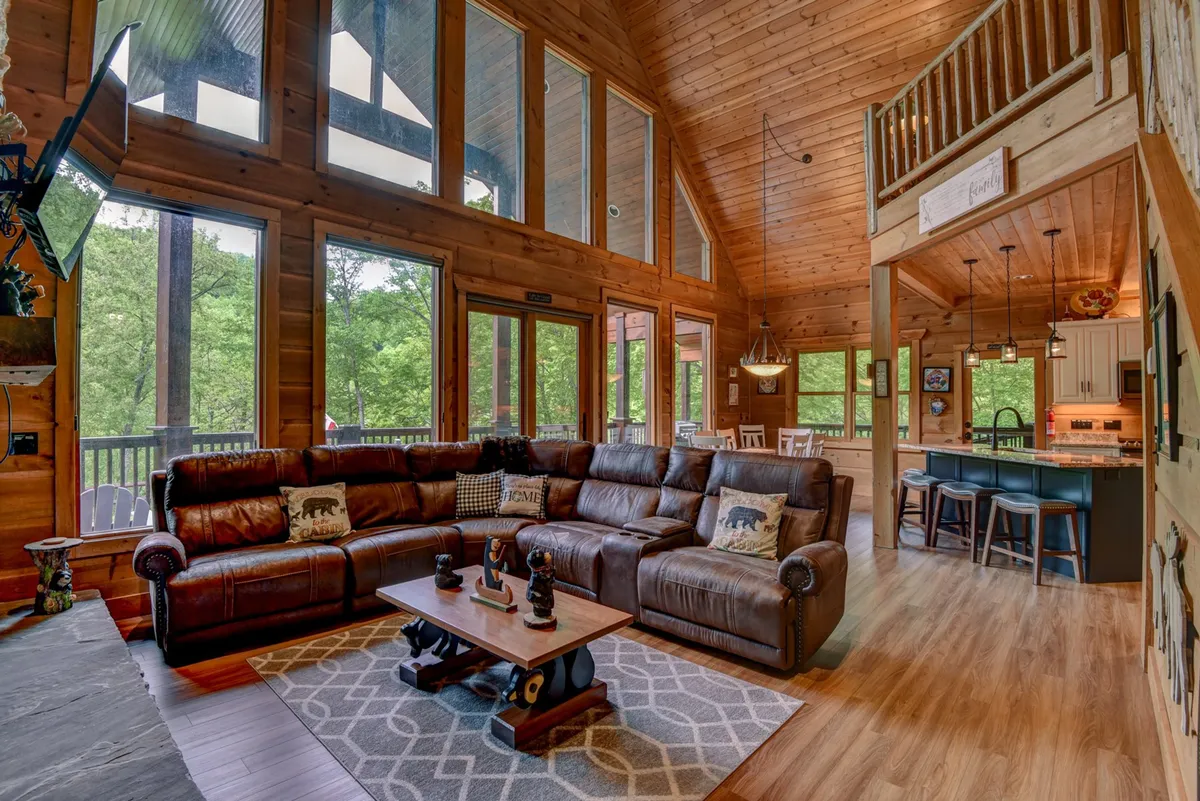 The spacious interior living area features a large, comfortable sectional sofa perfect for lounging, with floor-to-ceiling windows that let in natural light and offer views to the outdoors. A wooden dining table is nearby, enhancing the communal, homey atmosphere.