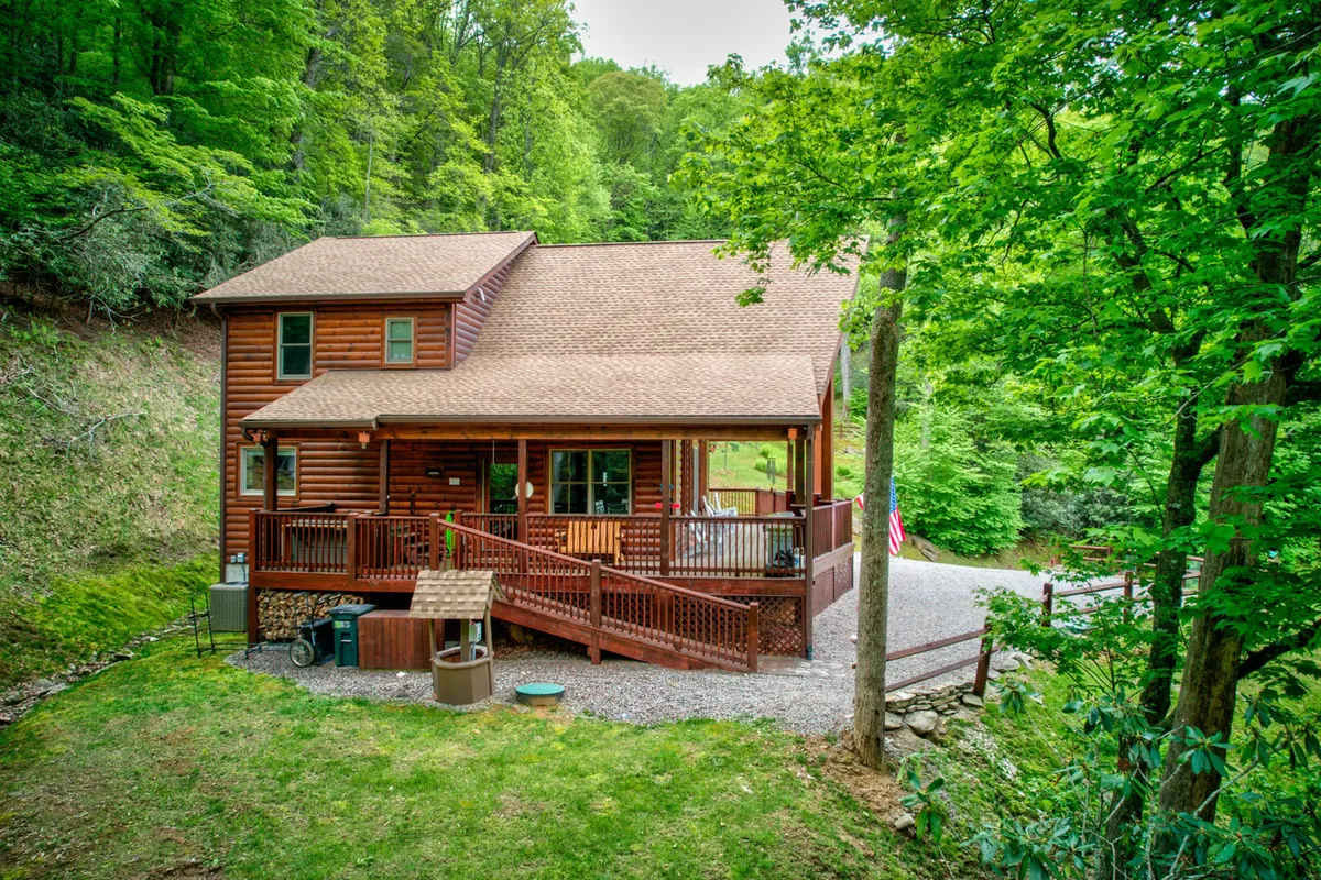 The exterior of the log cabin with a wooden deck, surrounded by trees and a gravel driveway. The porch is inviting, offering a spot to relax outdoors or return from a nature walk with your dog.
