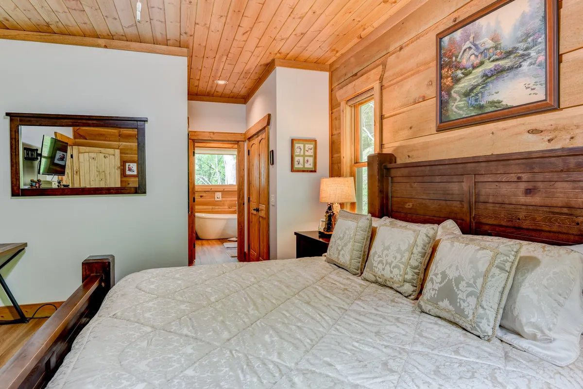 A cozy bedroom with a wooden headboard, decorative pillows, a bedside table with a lamp, and framed artwork above the bed. The inviting mood is enhanced by natural light, creating a welcoming space for restful sleep or quiet moments.