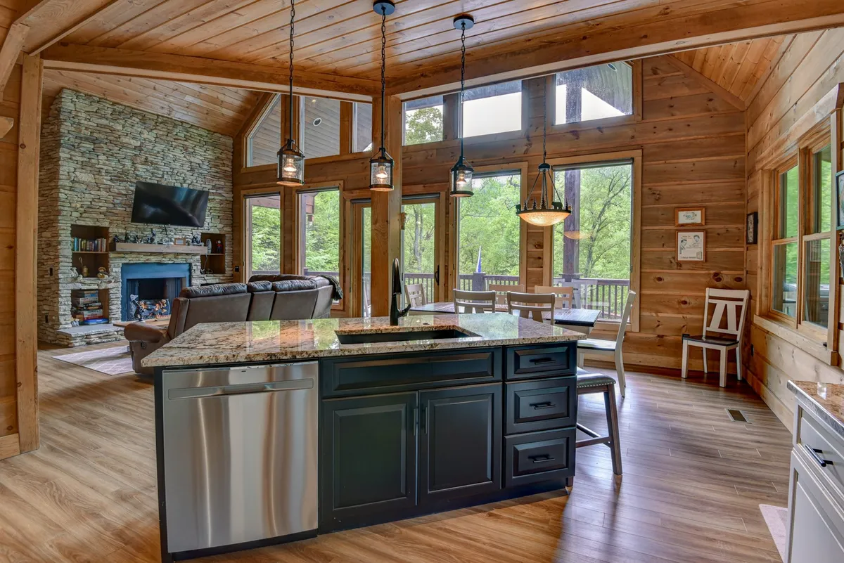 A spacious open-plan area featuring a granite kitchen island, a prominent stone fireplace, large windows with natural light, and exposed wooden beams. The living, kitchen, and dining spaces flow seamlessly together and are bathed in sunlight, creating a welcoming communal atmosphere.