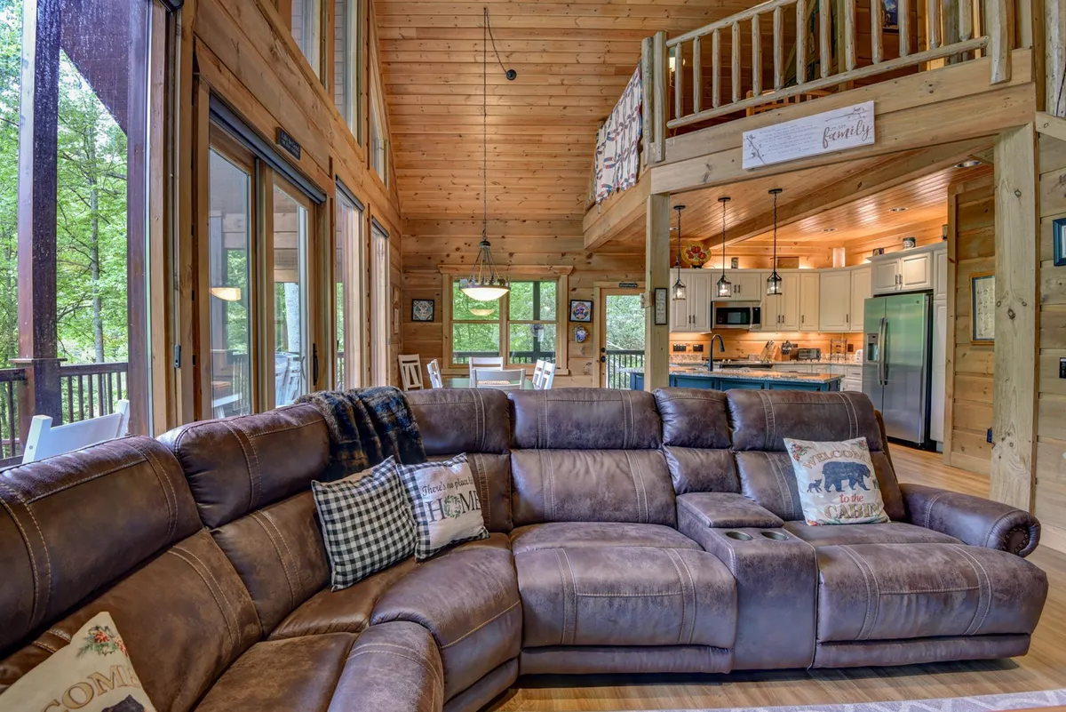 A wide-angle look at the large sectional sofa, wooden beams, kitchen, and dining table, all arranged in an open, sociable format. The setting emphasizes warmth, comfort, and group interaction.
