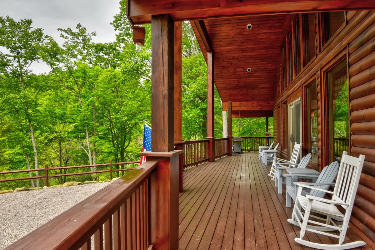A spacious wooden deck featuring classic rocking chairs overlooks a backdrop of lush green trees. The American flag adds a touch of welcoming charm, while the peaceful setting invites guests to relax and enjoy the fresh mountain air.